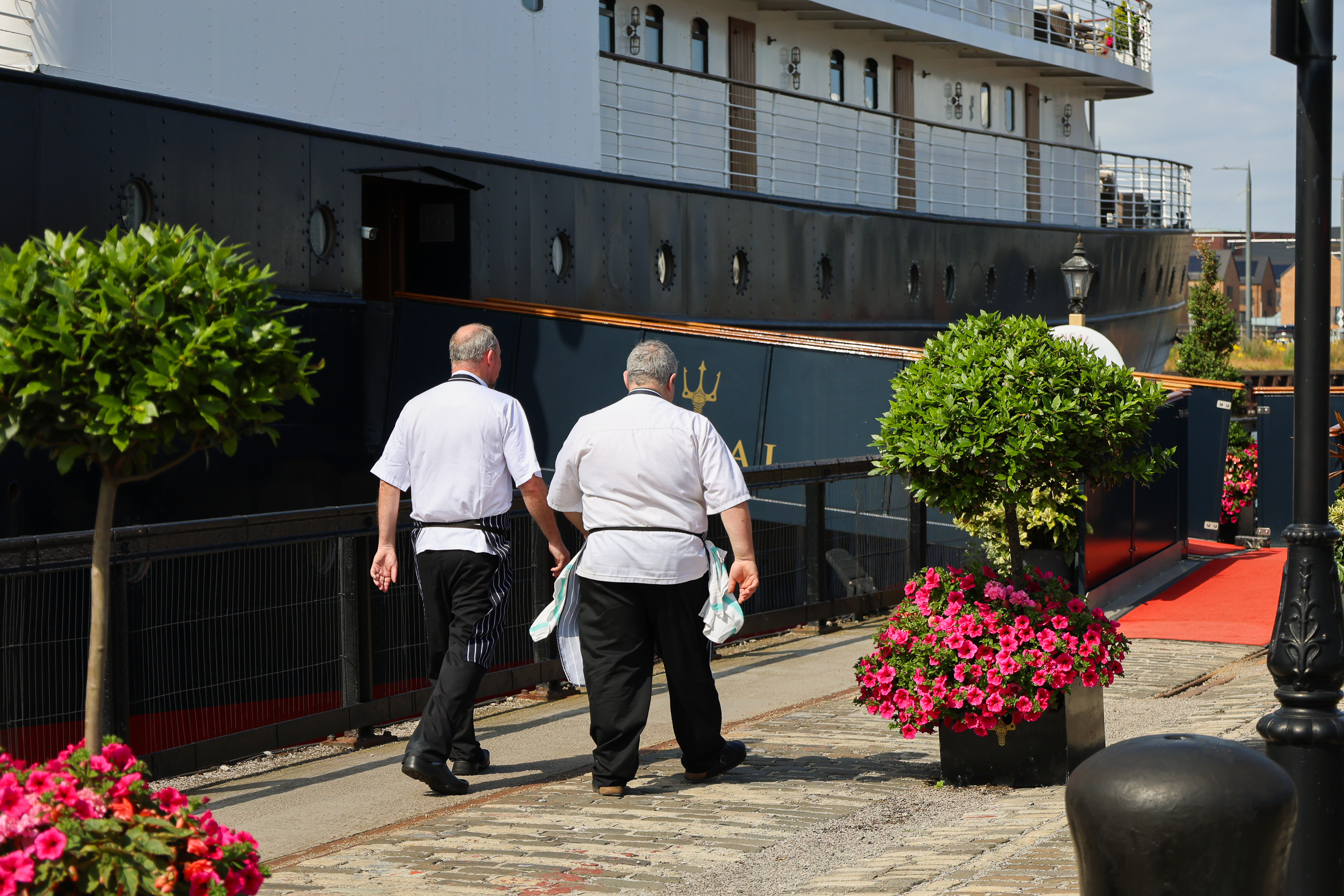 Two people walking beside a docked ship, with flowers lining the waterfront.