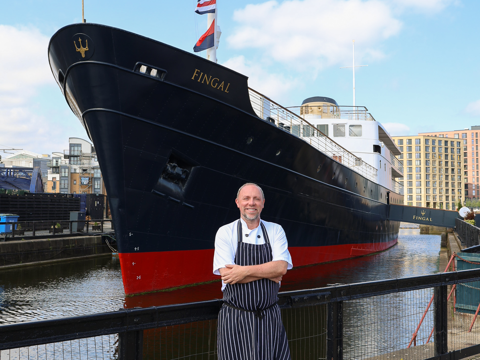Person in a chef’s uniform standing in front of a large ship at a harbor.