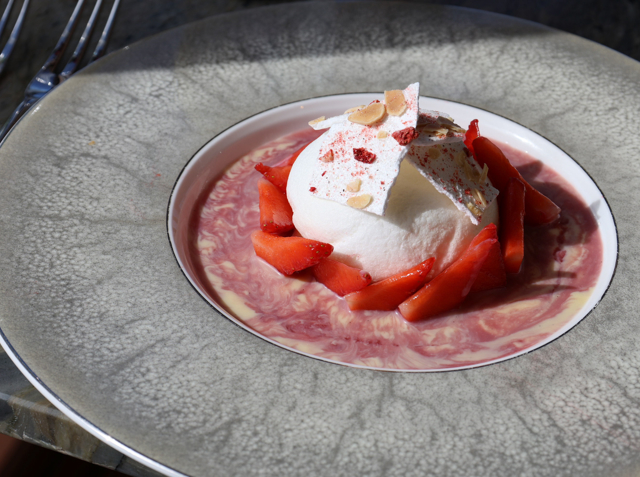 Poached meringue with Scottish strawberries arranged around it as crème anglaise is poured into the bowl, presented on a textured grey plate with a glass of lemon water beside it.