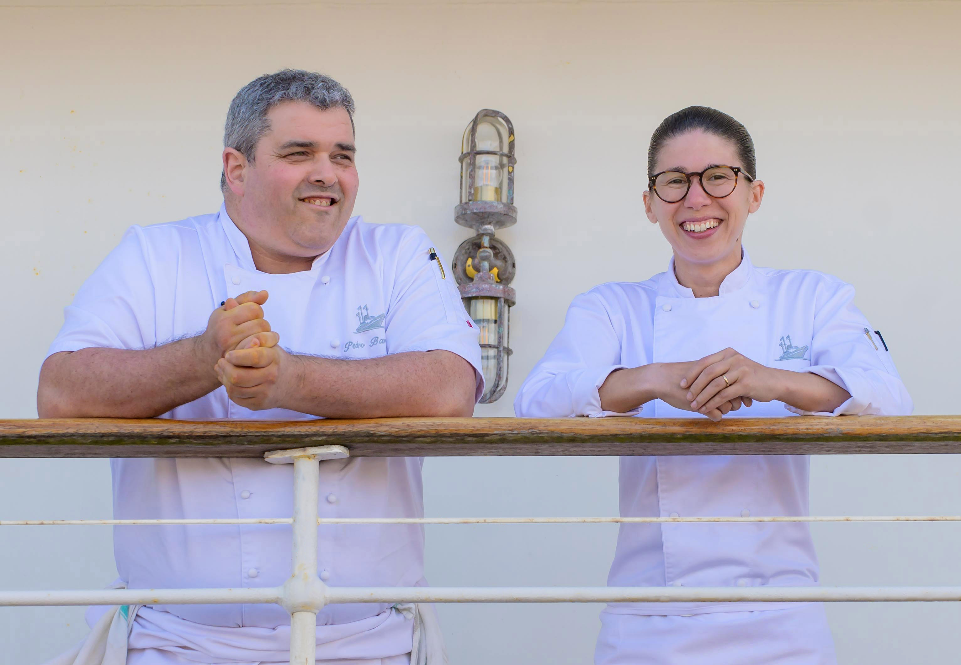 two chefs leaning over a ship railing smiling 