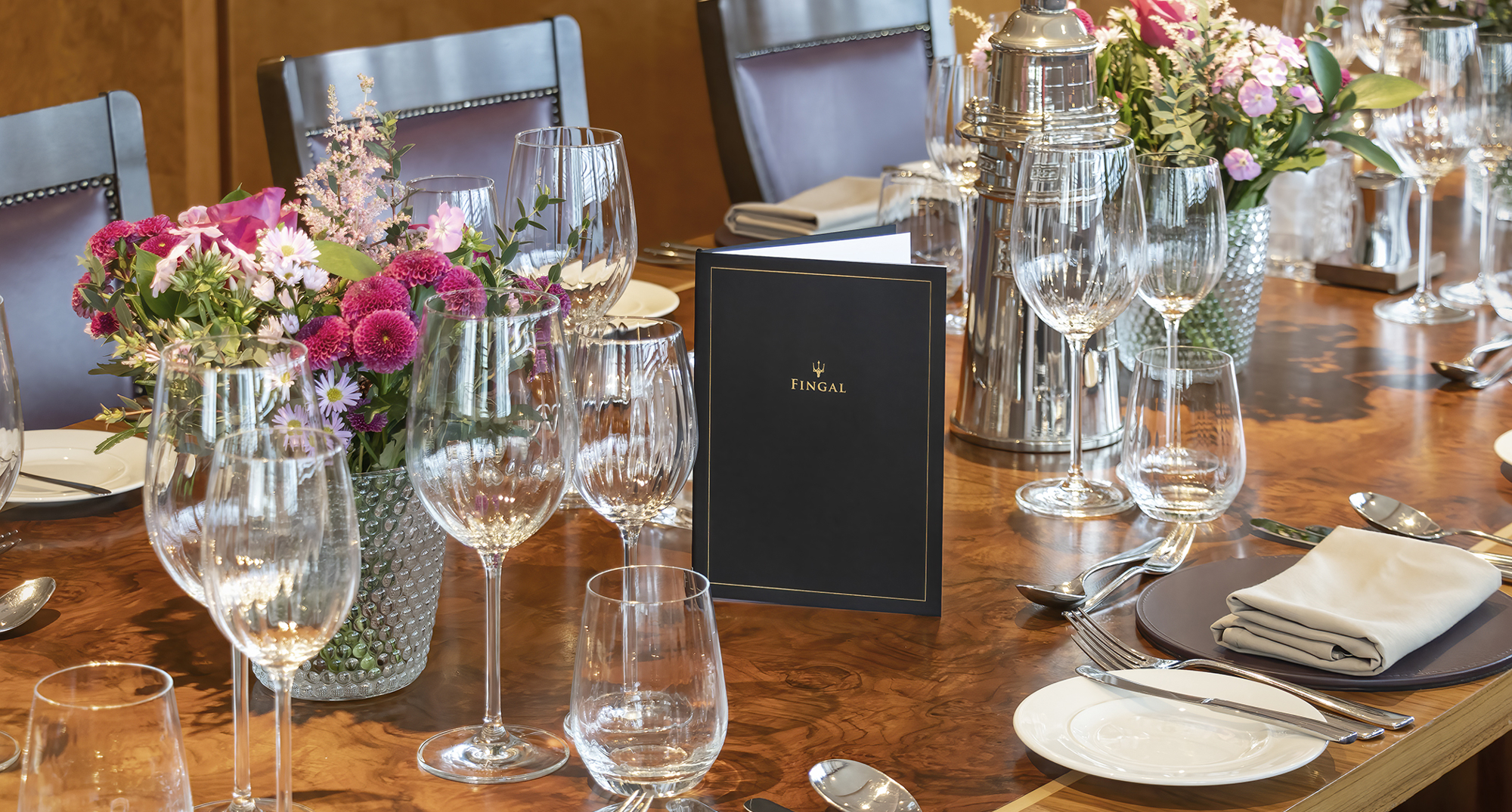 A table in Fingal's Bridge laid for dinner. There is china, glasses, cutlery, floral displays and menus on the beautiful wood table, and brow leather-backed chairs. 