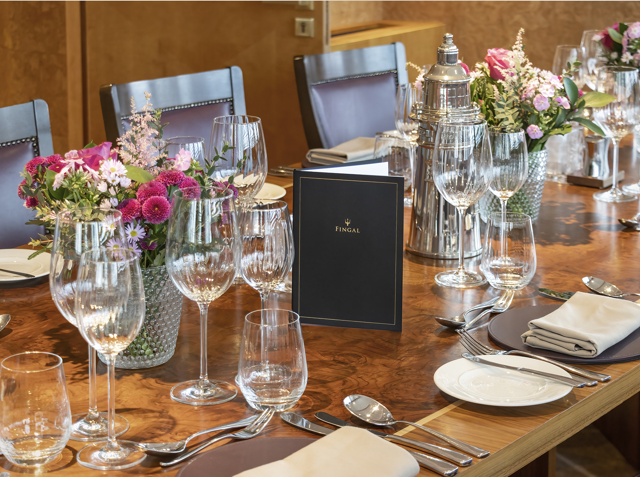 A table in Fingal's Bridge laid for dinner. There is china, glasses, cutlery, floral displays and menus on the beautiful wood table, and brow leather-backed chairs. 