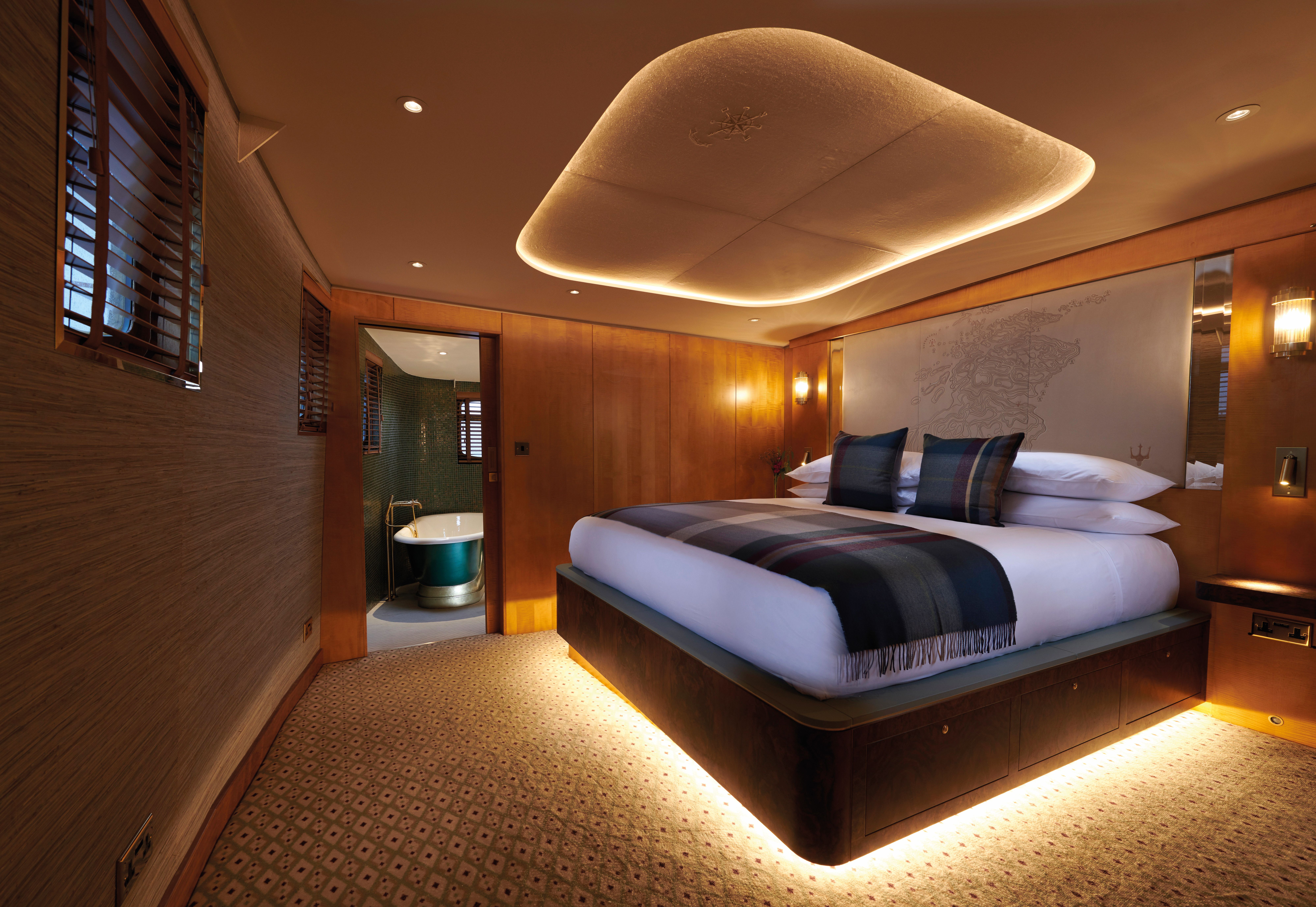 Modern bedroom with a large bed, soft wood‑panelled walls, and warm ambient lighting leading into a bathroom with a freestanding tub.