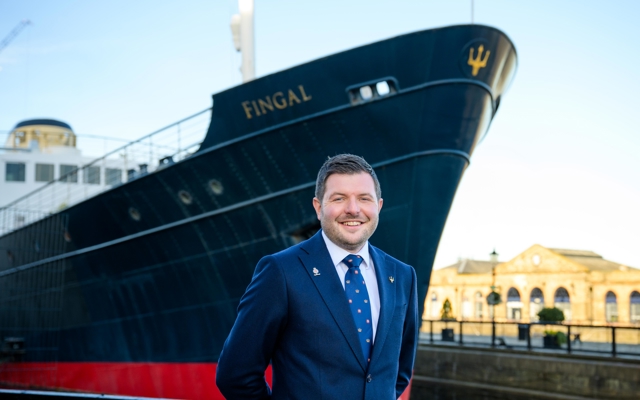 A man wearing a suit stands smiling at the bow of Fingal Hotel.