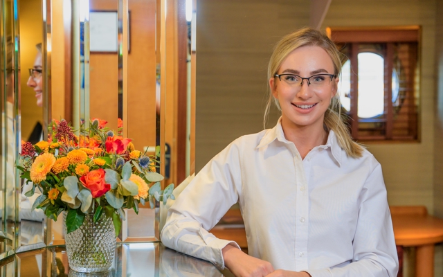 A woman wearing a white shirt is standing smiling next to a mirror in a cabin at Fingal Hotel.