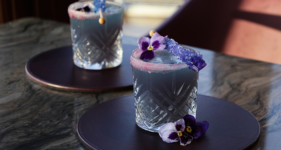 Purple coloured cocktail called Rare Gem which has a sugar cane crystal stick across the top of the glass. 