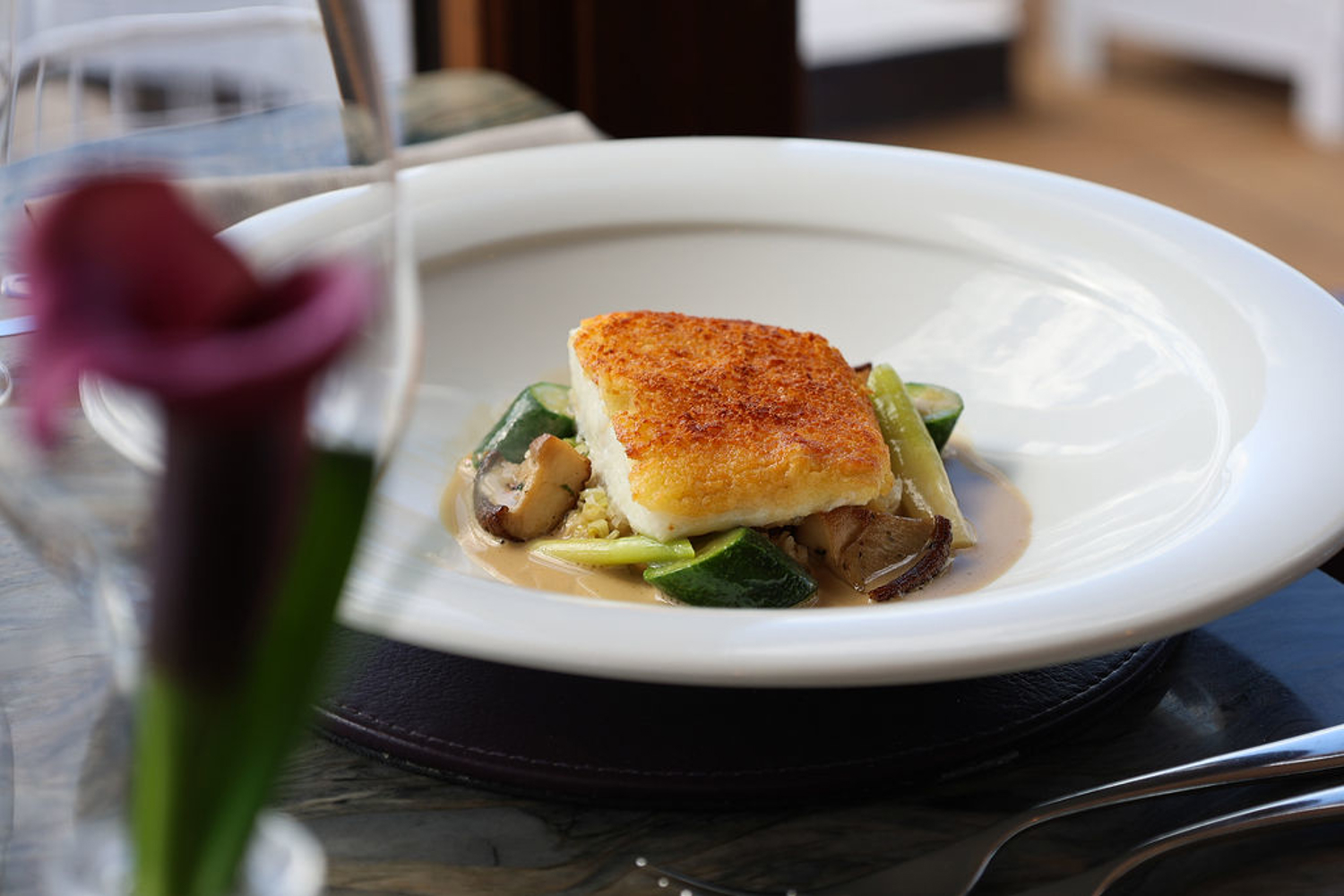 A seared piece of fish with a golden crust sits on a bed of vegetables and a creamy sauce, presented on a wide‑rimmed white plate in a fine‑dining style.