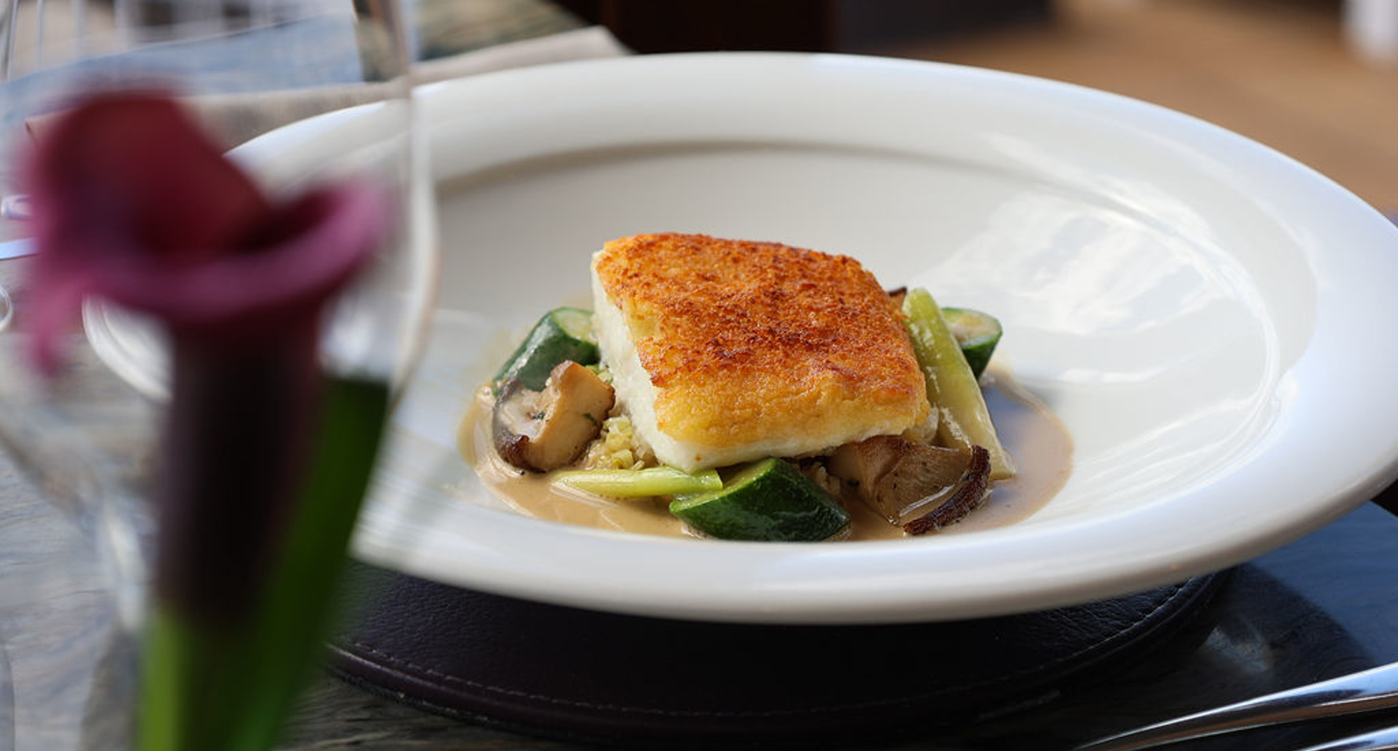 A seared piece of fish with a golden crust sits on a bed of vegetables and a creamy sauce, presented on a wide‑rimmed white plate in a fine‑dining style.
