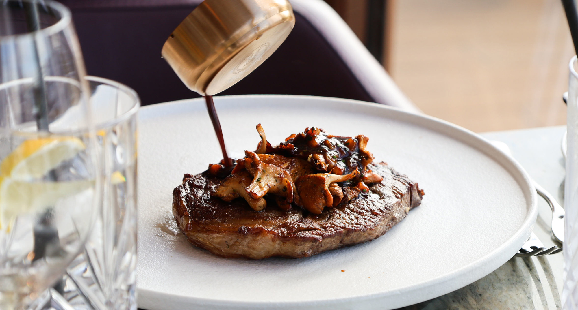 A cooked steak topped with sautéed Scottish mushrooms as a hand pours dark sauce from a small copper pan over the dish, set on a white plate in a fine‑dining setting.