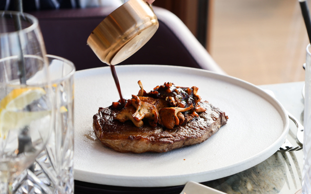A cooked steak topped with sautéed Scottish mushrooms as a hand pours dark sauce from a small copper pan over the dish, set on a white plate in a fine‑dining setting.