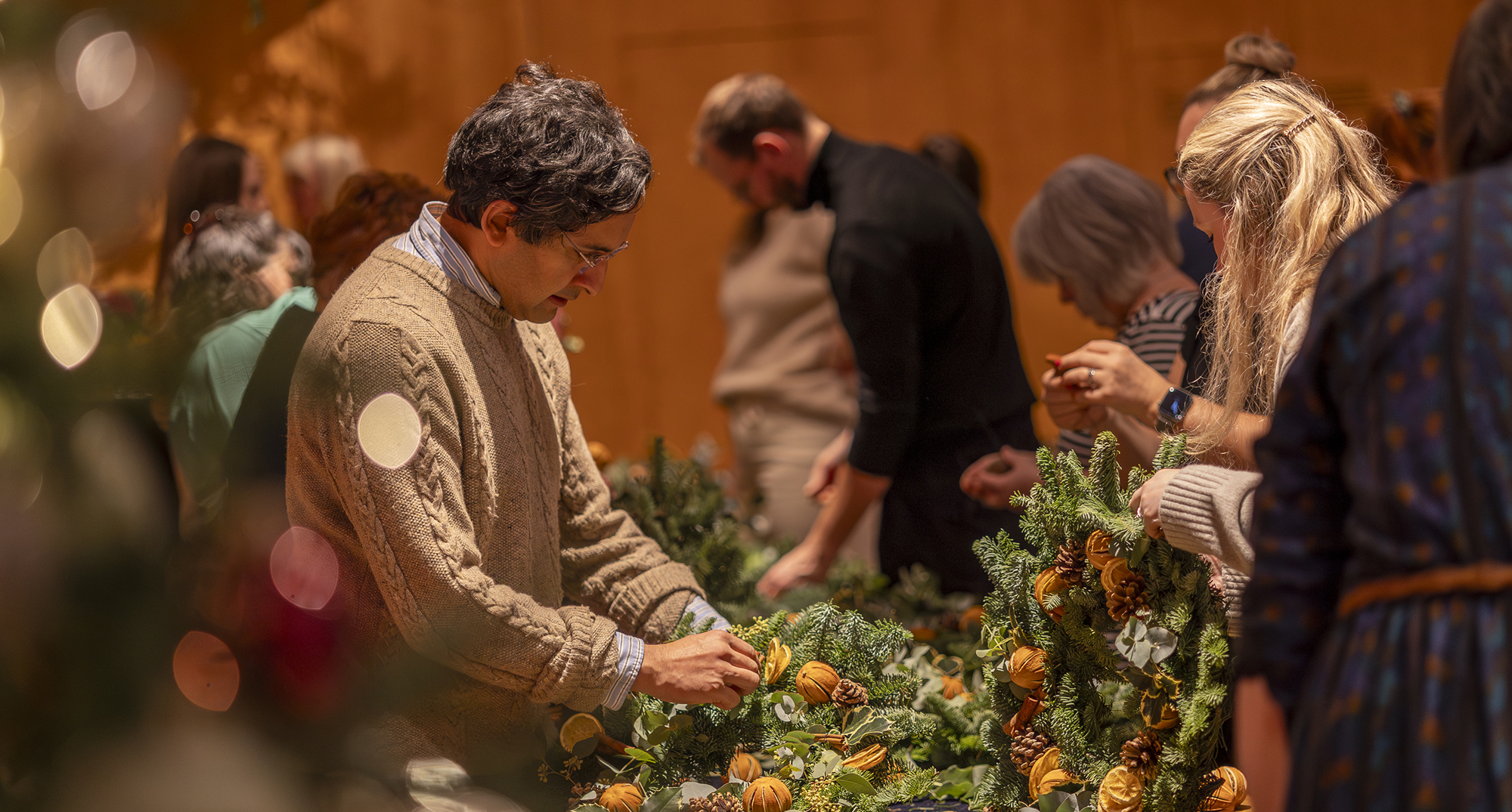 A group of people gathered around tables creating holiday wreaths with pine branches, dried oranges, and greenery.