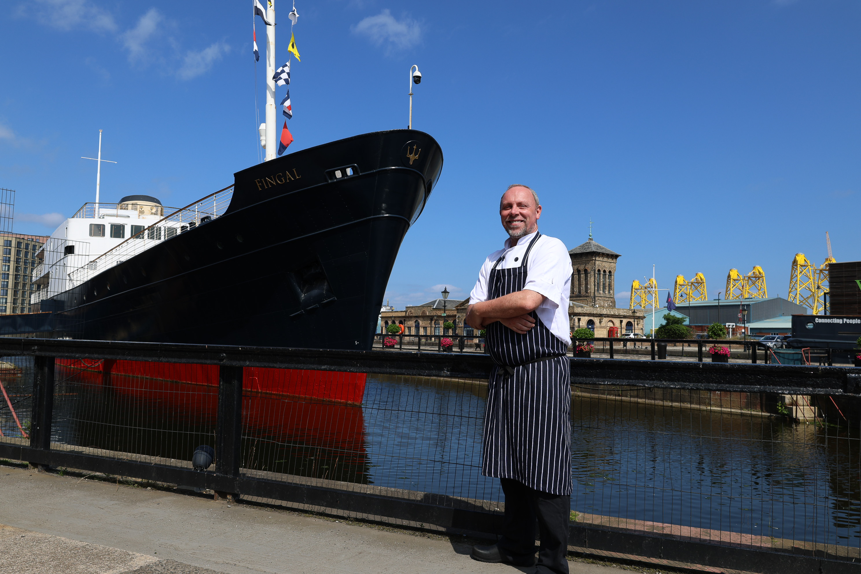 Person in a chef’s uniform standing beside a large ship in a harbor.