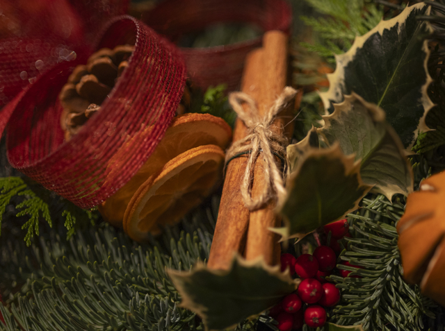 A close‑up of cinnamon sticks, dried oranges, pinecones, and holly arranged with red ribbon and evergreen foliage.