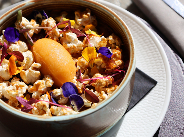 Acquerello rice pudding topped with salted caramel popcorn, edible flowers and a scoop of apricot sorbet, served in a ceramic bowl on a white plate with sunlight highlighting the textures.