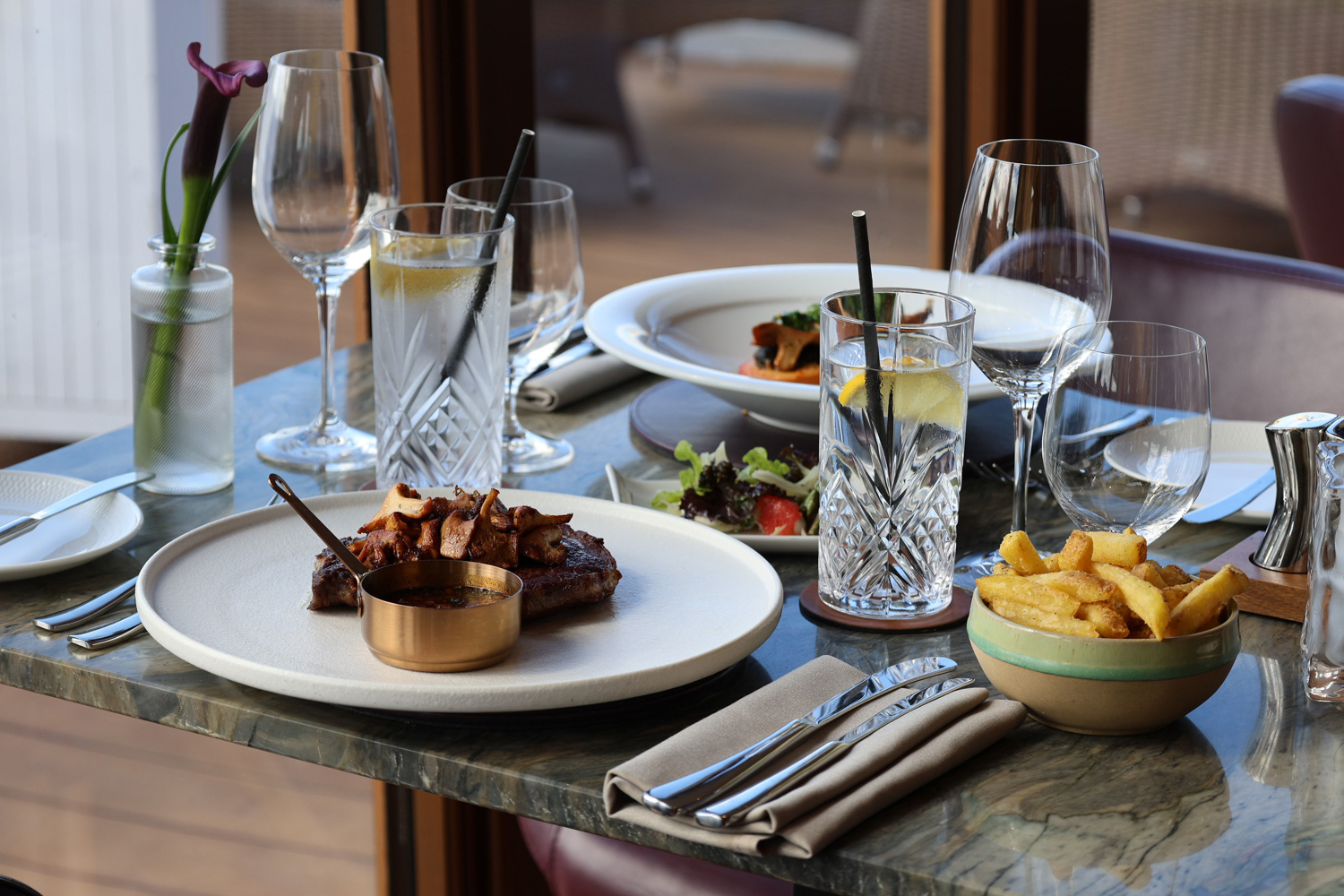 A restaurant table set with a grilled meat dish served with garnish and a small copper sauce pot, alongside a salad of greens and tomatoes, a bowl of thick‑cut fries, lemon‑garnished water glasses, wine glasses and a single purple flower in a small vase on a marble surface.
