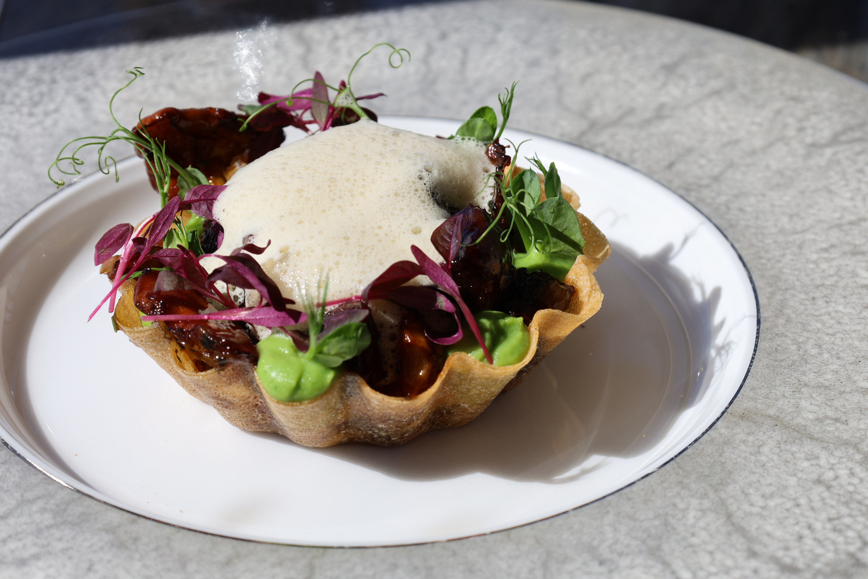 A delicate tart shell filled with green purée, micro herbs and vibrant garnishes, topped with a light foam and presented on a white plate with a decorative rim.