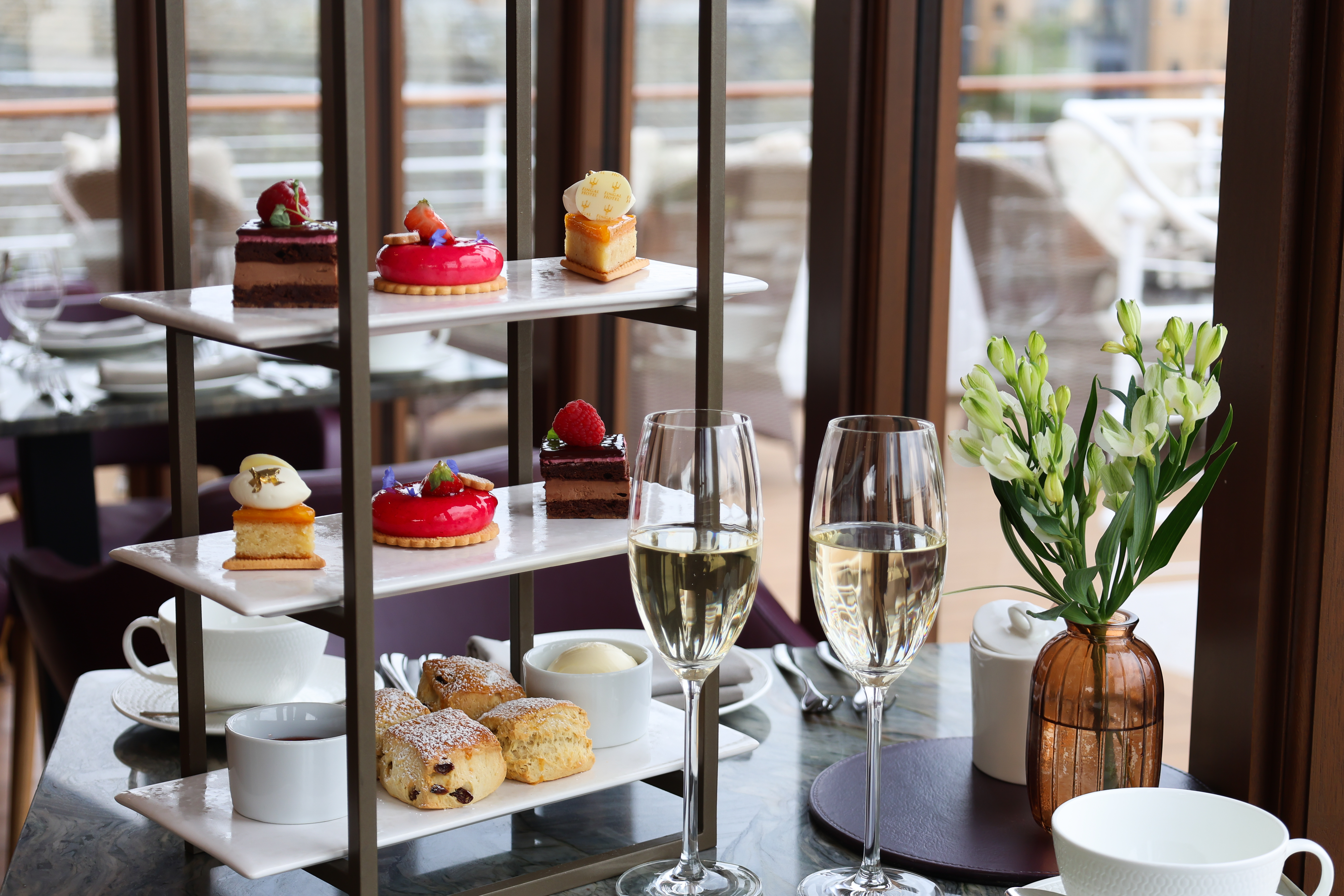 Three‑tier afternoon‑tea stand with pastries, tarts, and scones, served with sparkling wine in a bright dining room
