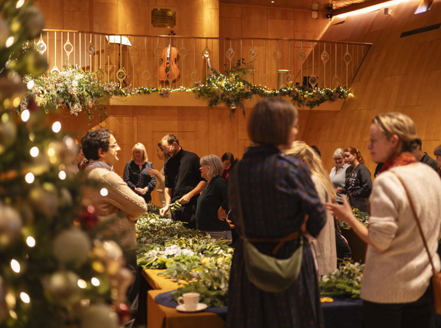 Guests gathered in a warmly lit space, crafting with greenery and wreath materials beside a decorated Christmas tree.