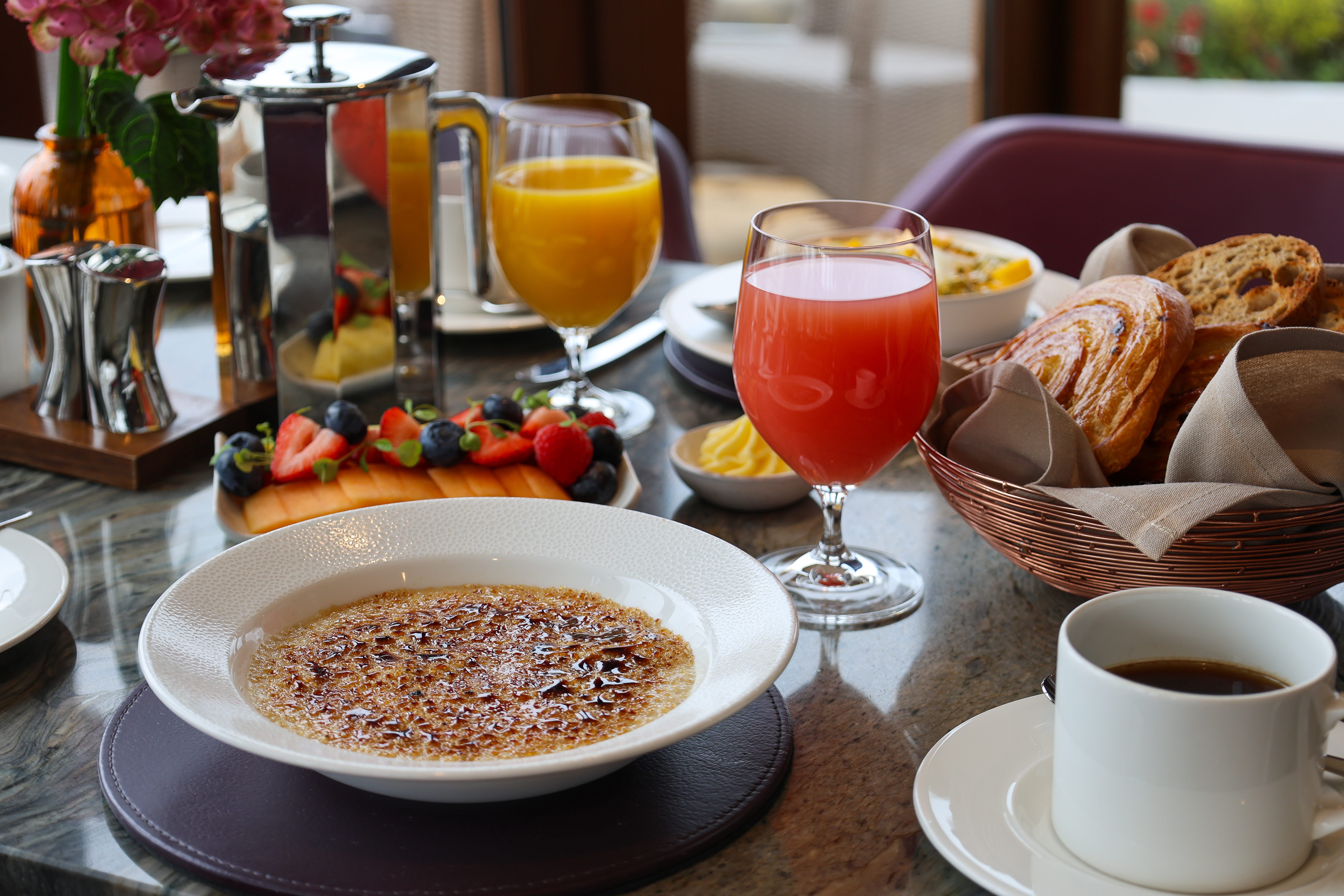 Breakfast spread with crème brûlée, pastries, coffee, fresh fruit, and glasses of juice on a café table.