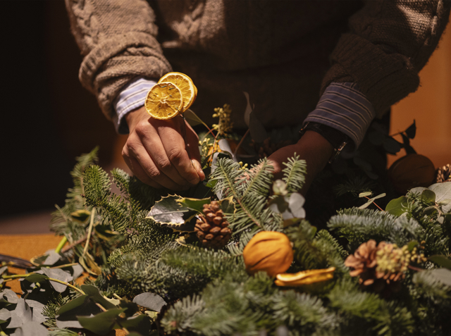 A person arranging dried orange slices into a festive wreath of evergreens, pinecones, and natural décor.