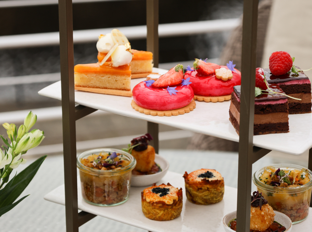 Tiered stand with colourful pastries and savoury jars, set against a blurred waterside backdrop.