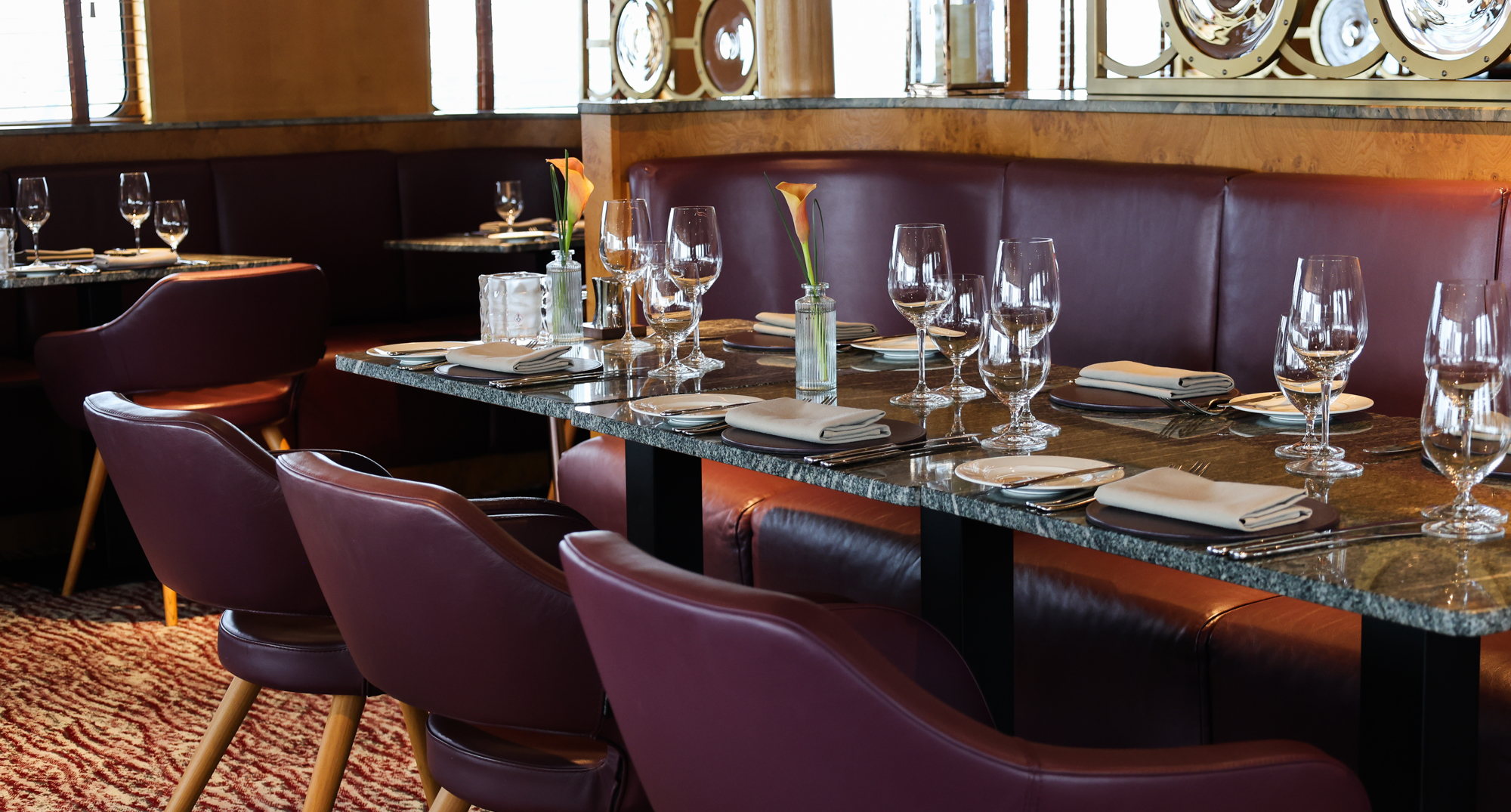 Long marble table set with folded napkins, glassware, and a single orange calla lily, surrounded by burgundy seating.