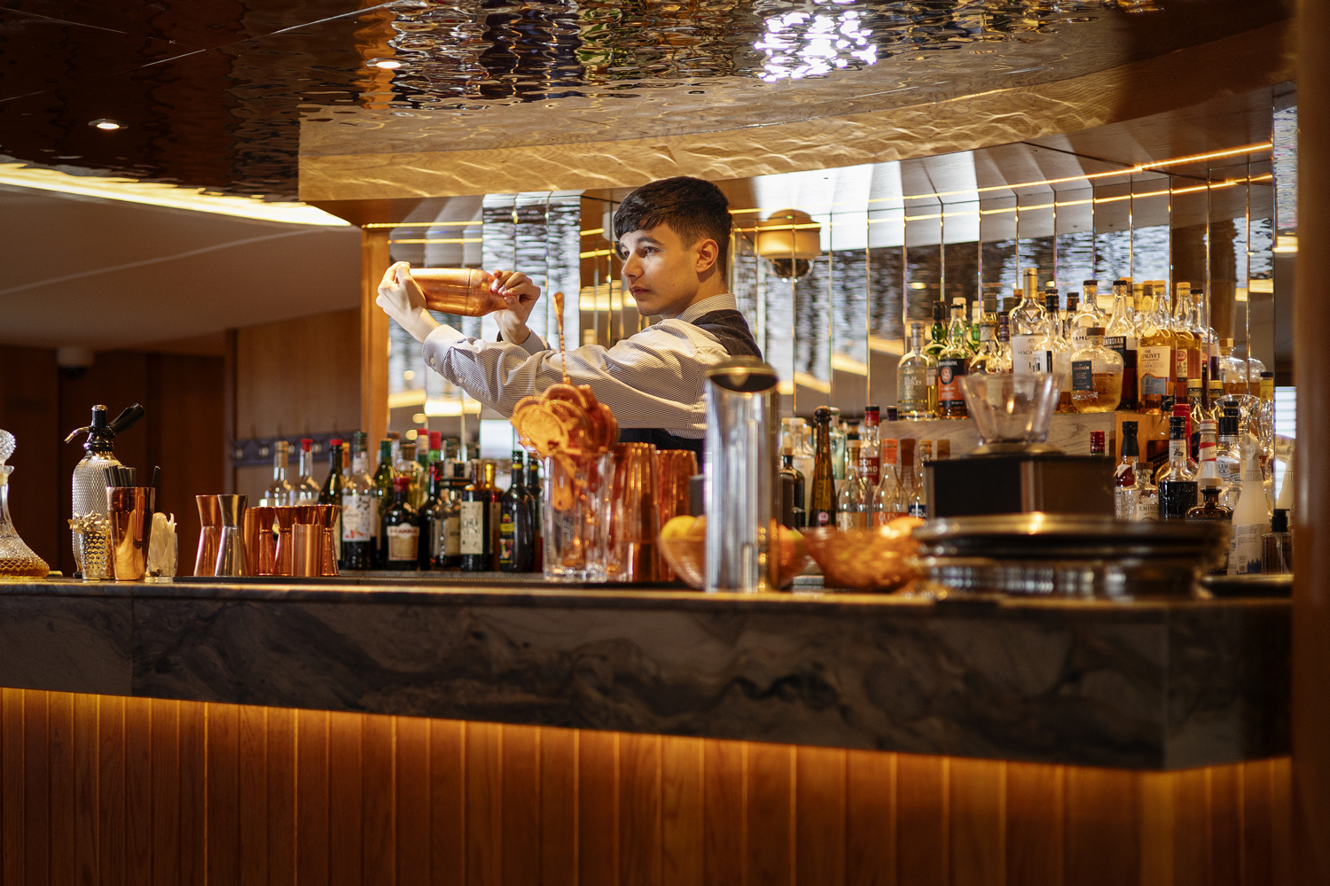 Bartender shaking a cocktail behind a well‑lit bar stocked with bottles and copper barware.