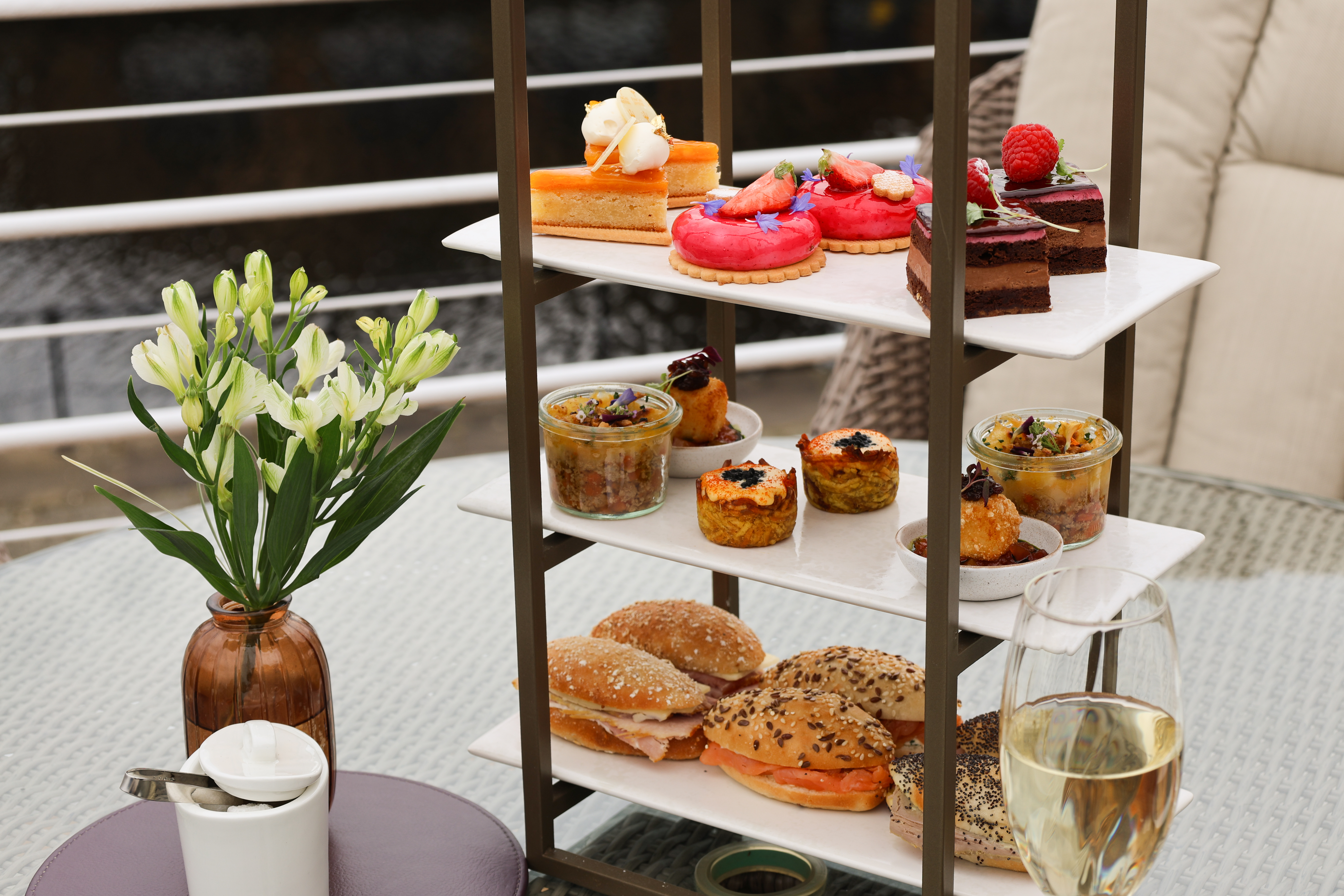 Three‑tier afternoon tea stand with colourful pastries, savoury jars, and sandwiches on an outdoor waterside table.