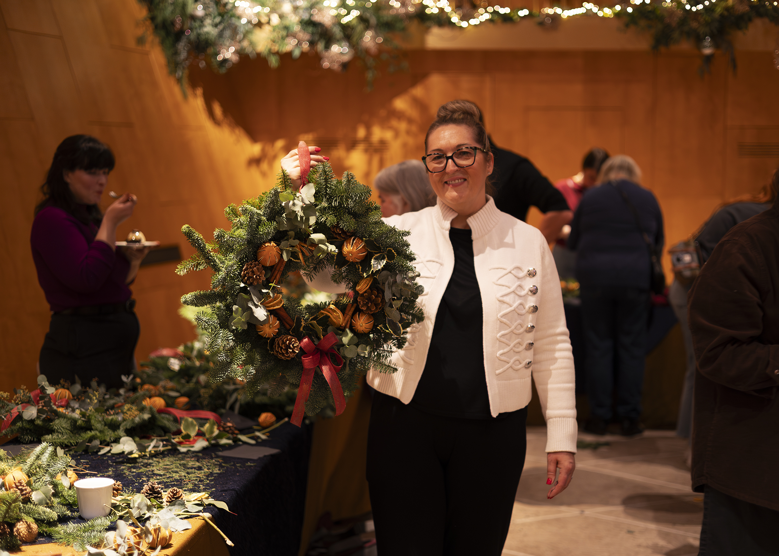 A person holding a decorated holiday wreath made with evergreens, pinecones, dried oranges, and red ribbon at a cosy workshop.