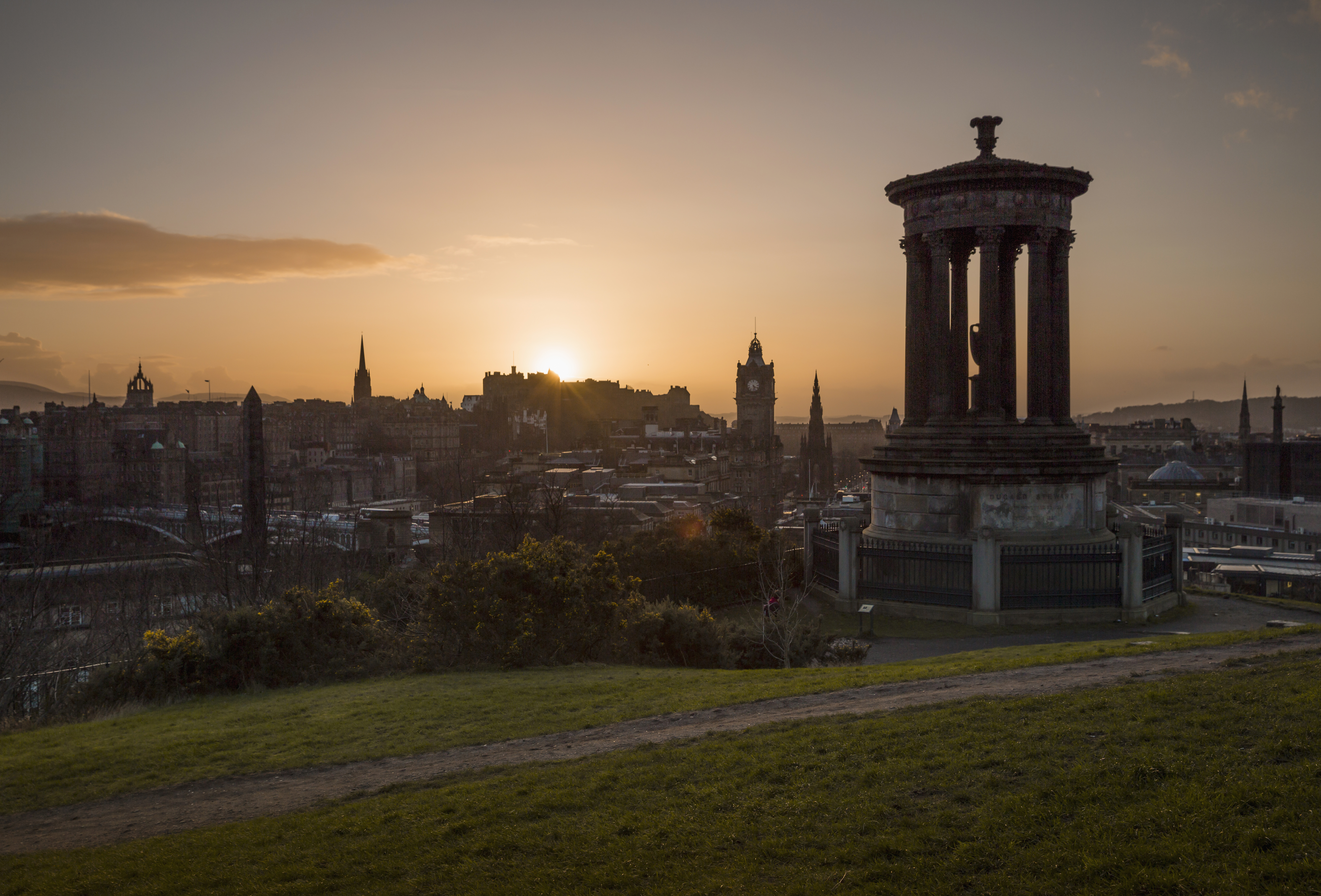 Sunset over Calton Hill in Edinburgh. A tall monument is in the foreground and the city skyline is in the background. 
