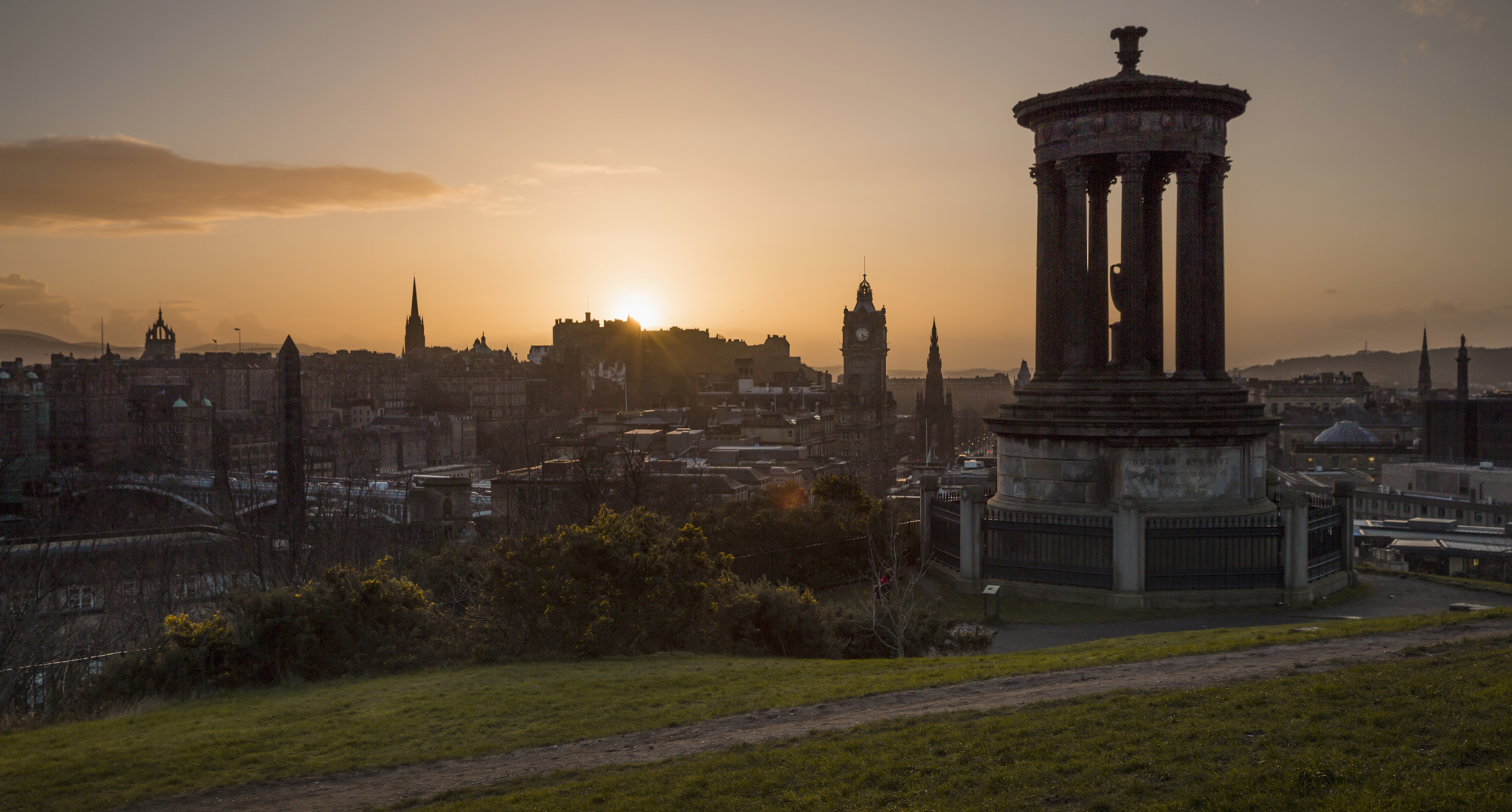 Sunset over Calton Hill in Edinburgh. A tall monument is in the foreground and the city skyline is in the background. 