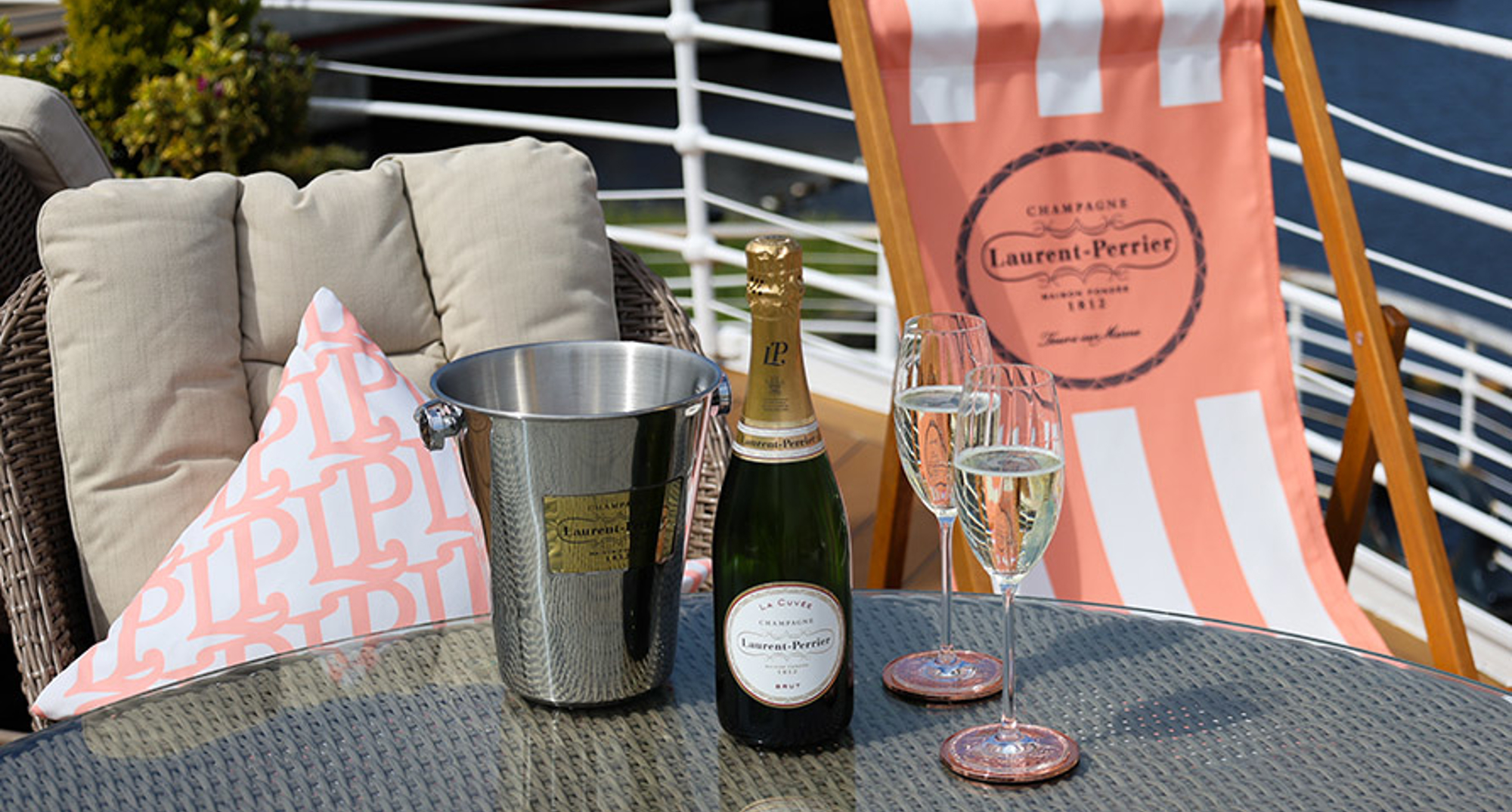 A pink and white striped deck chair next to a table containing a bottle of Champagne, silver ice bucket and glass flutes. 