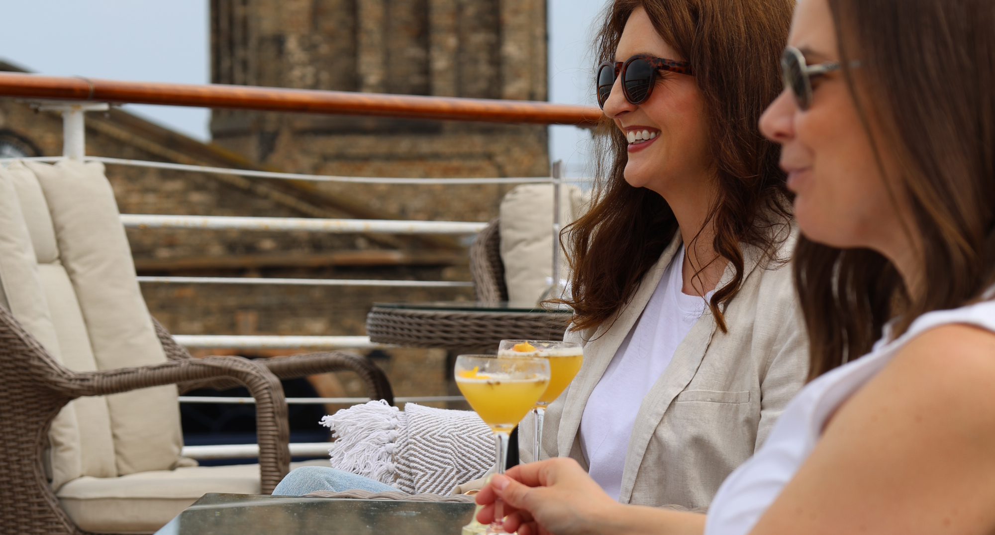 Two people sit on wicker chairs outdoors, each holding an orange‑coloured cocktail in a stemmed glass. They wear sunglasses and light clothing, relaxing at a table on a sunny deck with a stone building rising in the background.