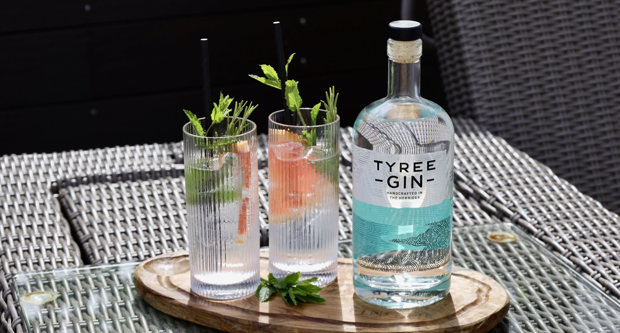A bottle and two glasses of gin on a patio in the sunshine.