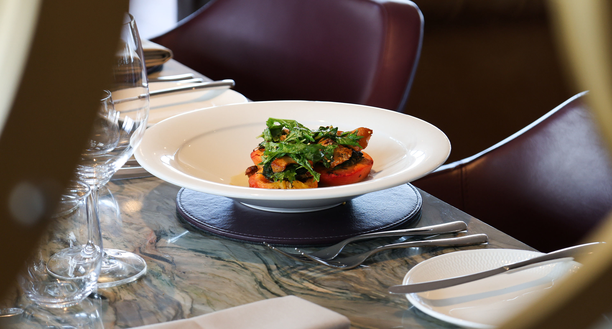 A white bowl holds a dish of roast peach with nasturtium, roquette and foraged Scottish mushrooms, set on a marble‑patterned table with wine glasses, cutlery and a folded napkin creating an elegant dining scene.