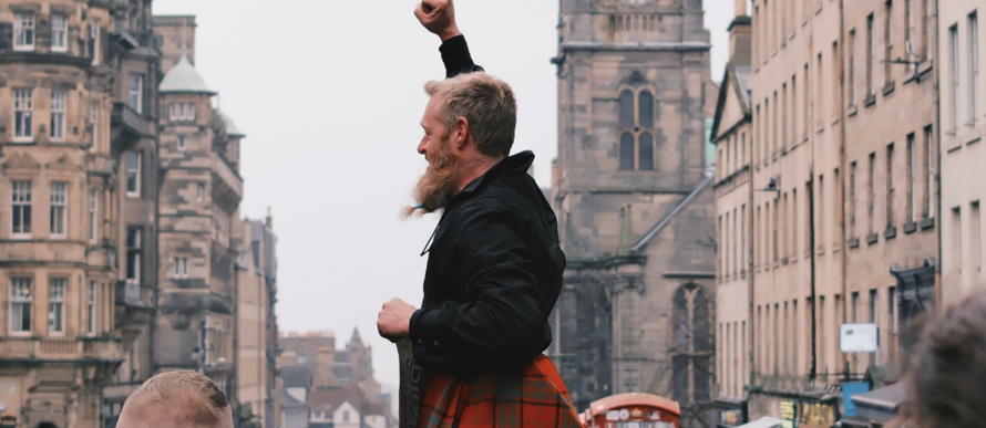 Person wearing a red tartan kilt stands above a crowd on a city street, raising a fist, with historic buildings and a clock tower in the background.