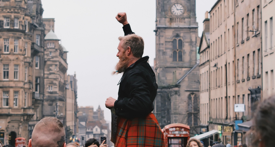 Person wearing a red tartan kilt stands above a crowd on a city street, raising a fist, with historic buildings and a clock tower in the background.