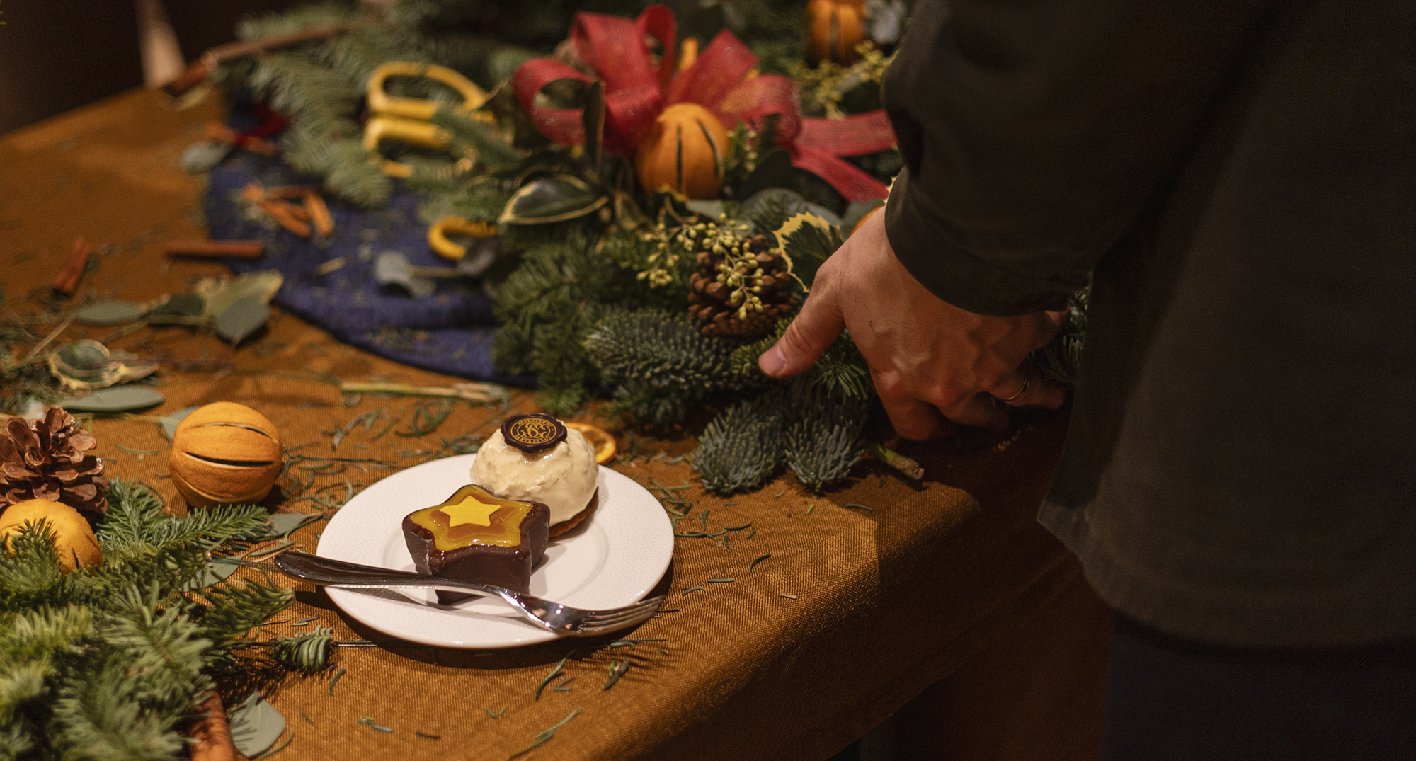 A hand adjusting a holiday greenery arrangement with pinecones, dried oranges, and red ribbon beside a plated dessert.