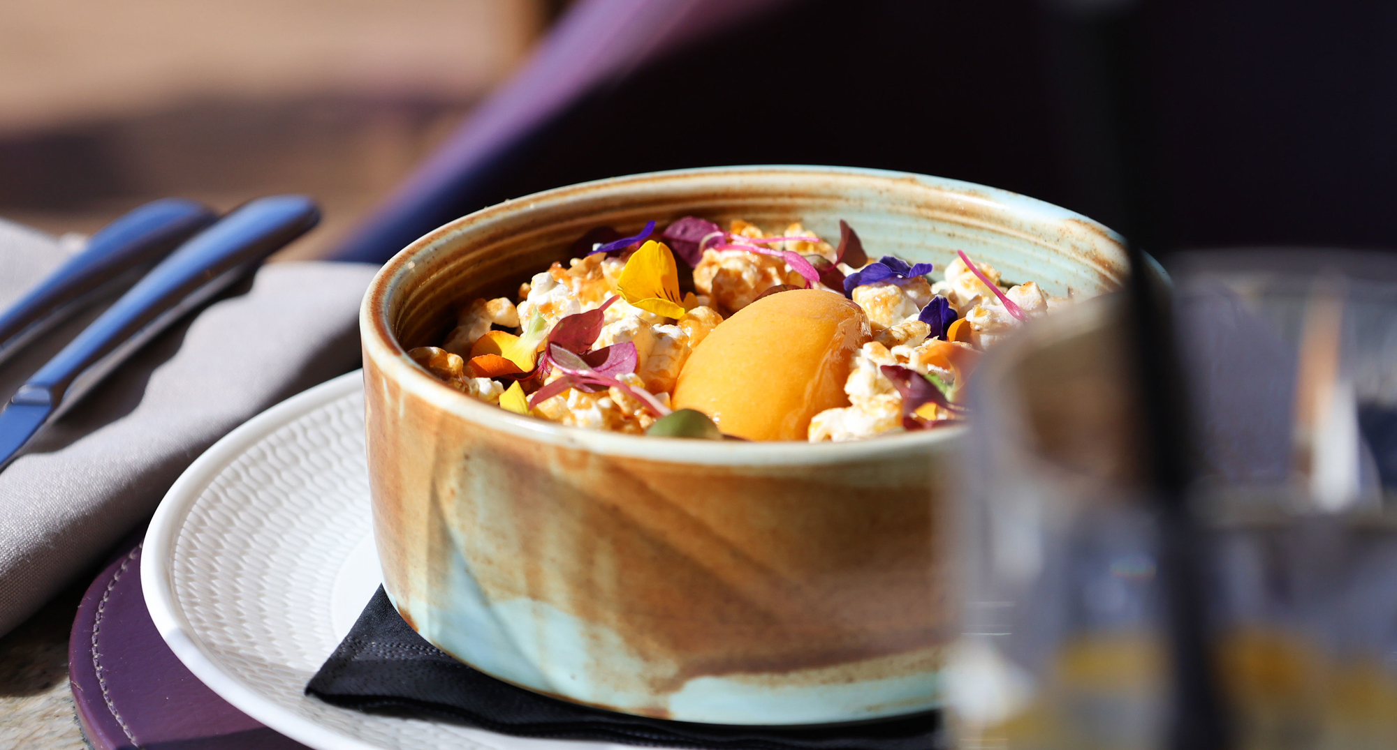 Acquerello rice pudding topped with salted caramel popcorn, edible flowers and a scoop of apricot sorbet, served in a ceramic bowl on a white plate with sunlight highlighting the textures.