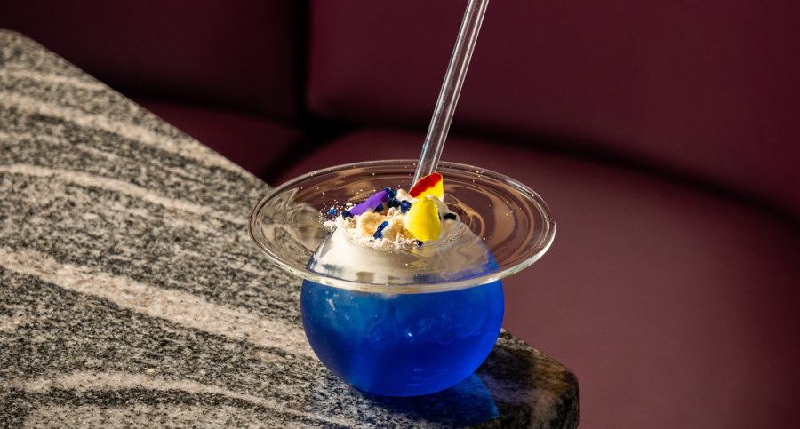 Blue cocktail with a citrus garnish and straw, served on a stone table against a burgundy booth backdrop.