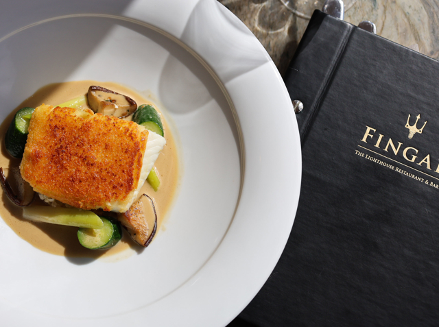 A golden‑seared piece of fish rests on Brussels sprouts and mushrooms with a light brown sauce, served in a wide‑rimmed white bowl beside a black menu embossed in gold with “FINGAL The Lighthouse Restaurant & Bar.”