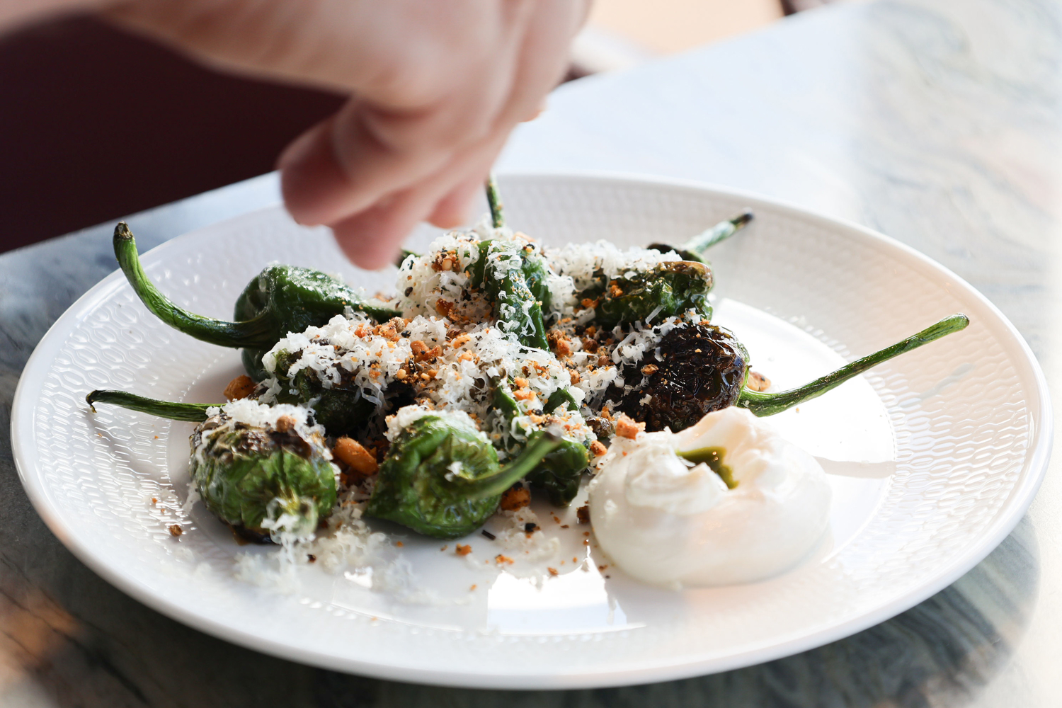 A plate of blistered green peppers topped with grated cheese and crushed seasoning, served with a dollop of creamy white sauce. A hand hovers above, sprinkling a finishing touch over the dish on a white textured plate set on a marble surface.