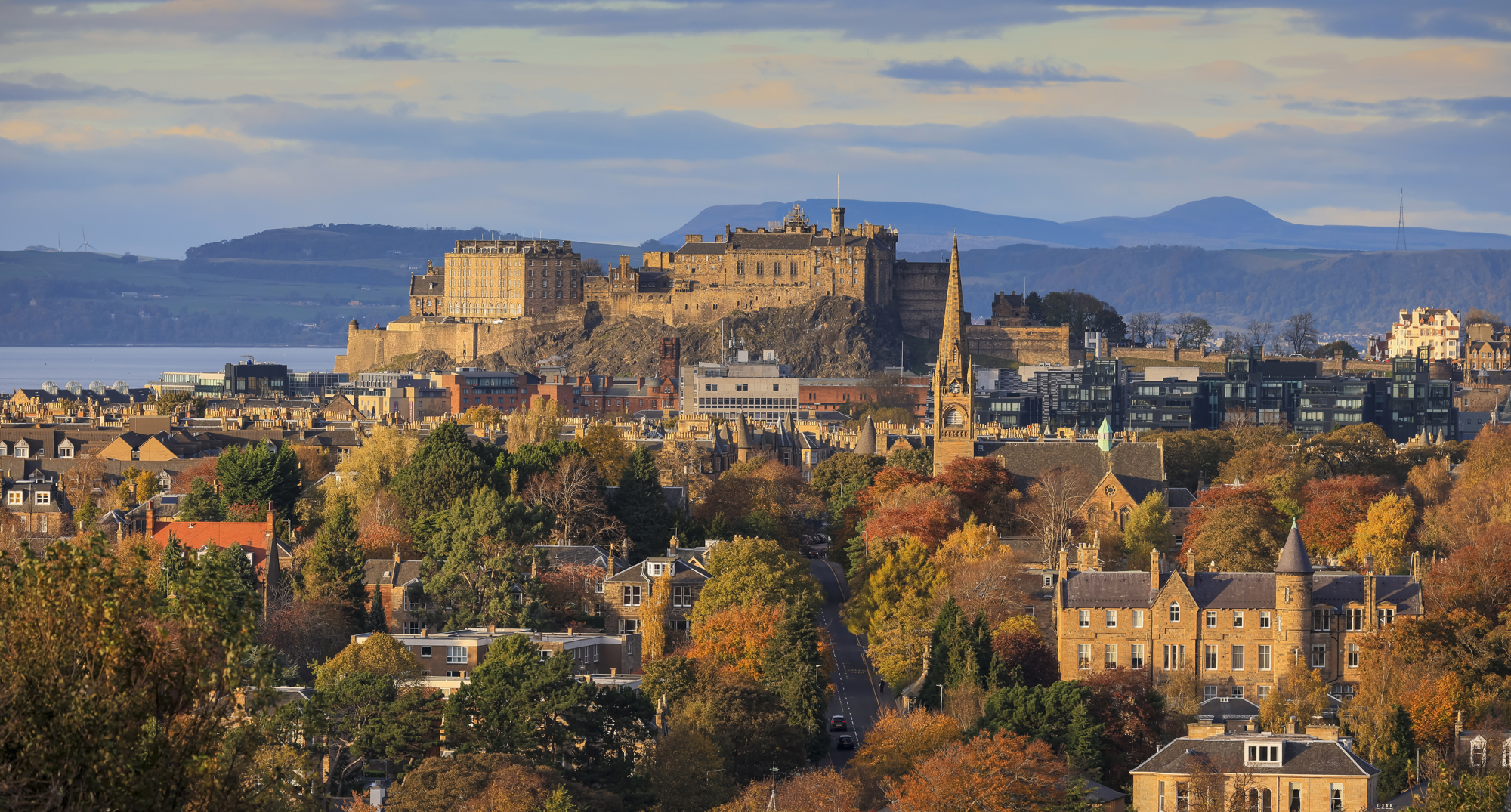 Edinburgh Castle and the city skyline on a bright day. The sun is beginning to set and the buildings are bathed in golden light. 