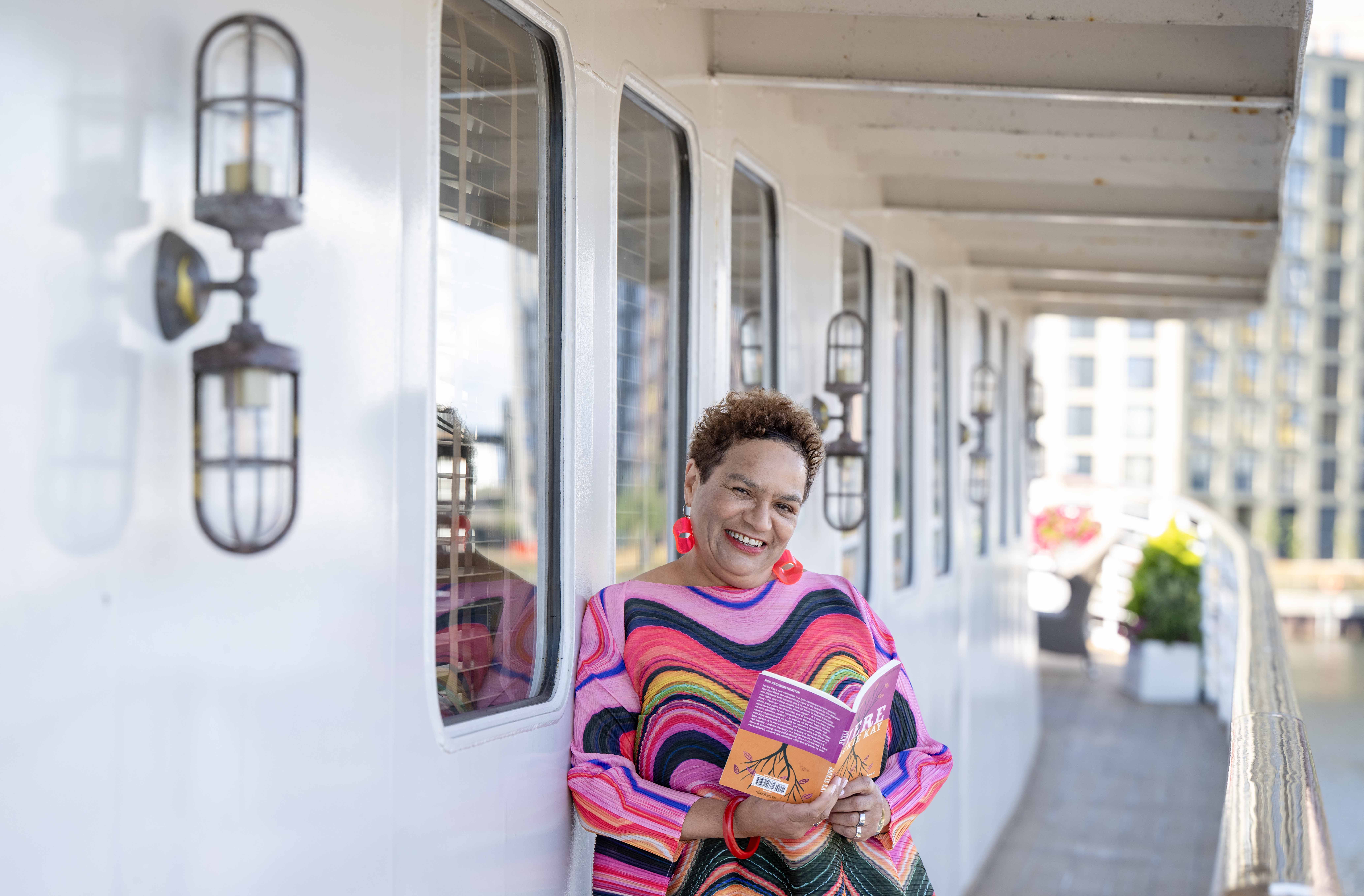 A woman in bright clothing on a ship deck with a book in her hands.