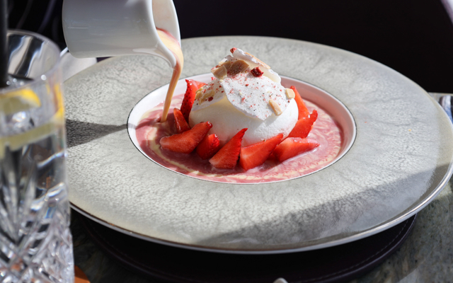 Poached meringue with Scottish strawberries arranged around it as crème anglaise is poured into the bowl, presented on a textured grey plate with a glass of lemon water beside it.