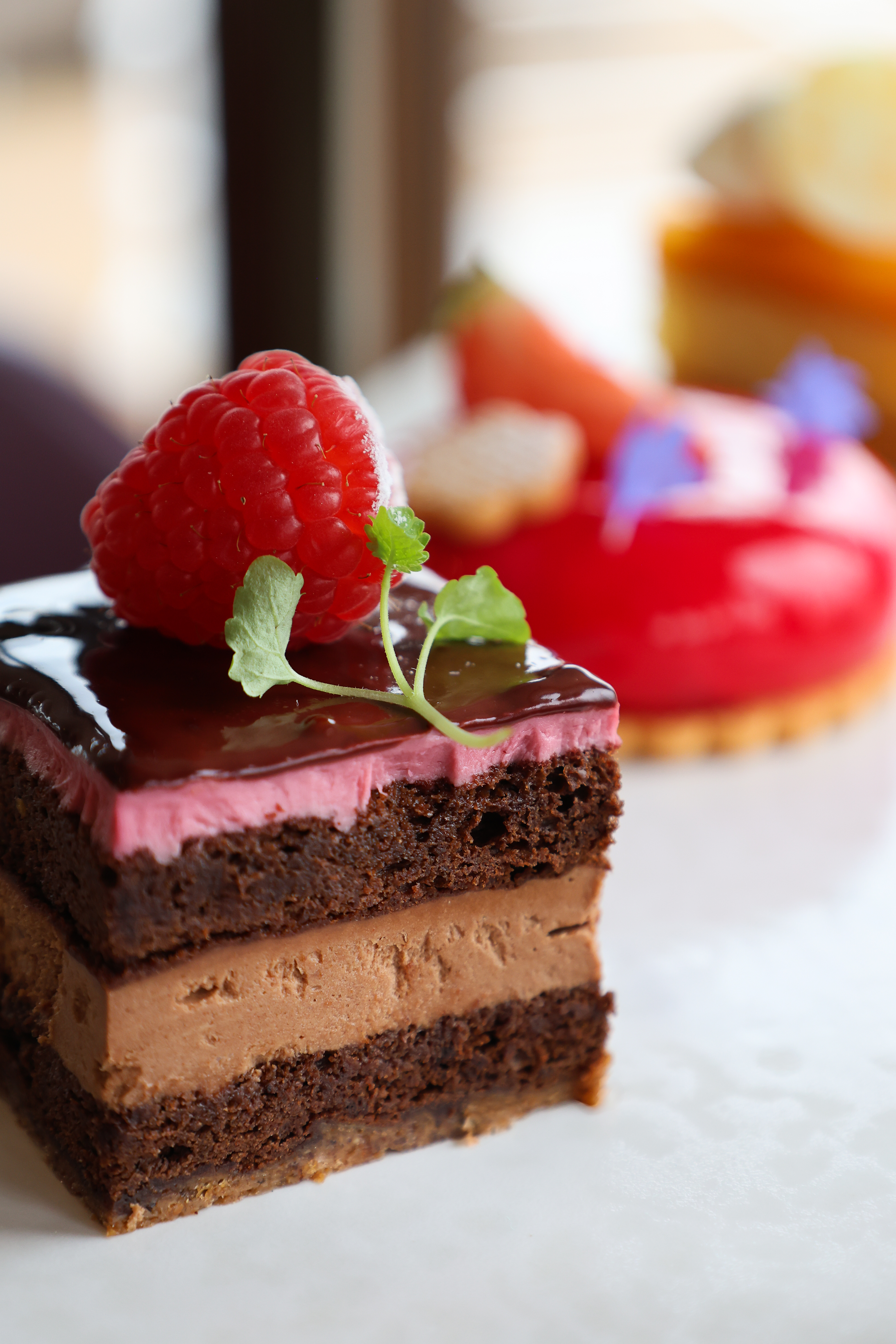 Layered chocolate mousse cake topped with a raspberry and mint, with colourful pastries blurred behind.