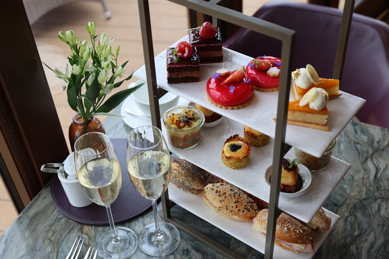 Three‑tier stand with colourful pastries, layered parfaits, and mini sandwiches, served with champagne in a restaurant.