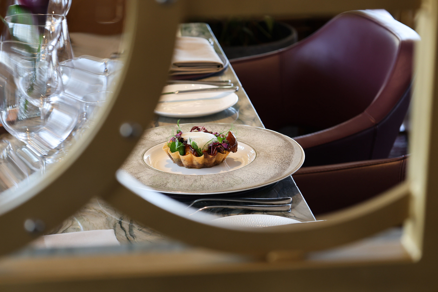 A fine‑dining table setting viewed through a circular frame, featuring a small tart topped with greens and garnishes on a white plate, with silverware, a folded napkin and glassware arranged neatly beside a maroon upholstered chair.